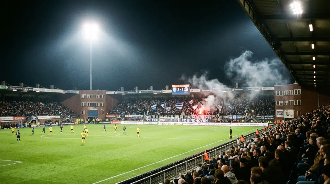 Klein Nederlands voetbalstadion met intieme sfeer tijdens een avondwedstrijd onder floodlights
